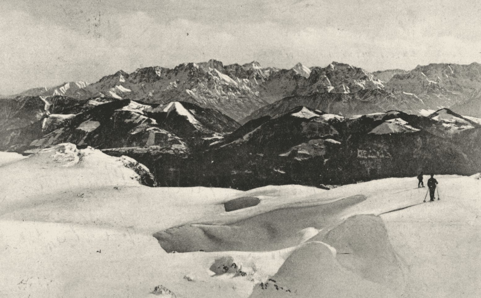 Two figures crossing deep snow below a row of peaks, 1912 postcard